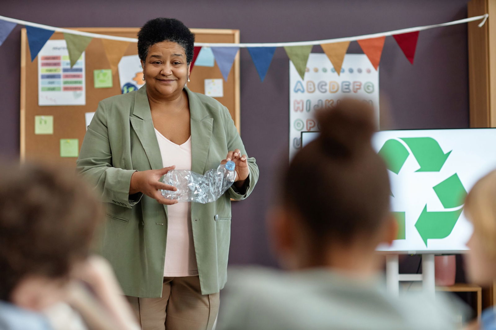Waist up portrait of smiling Black woman teaching little kids about recycling and waste sorting in preschool